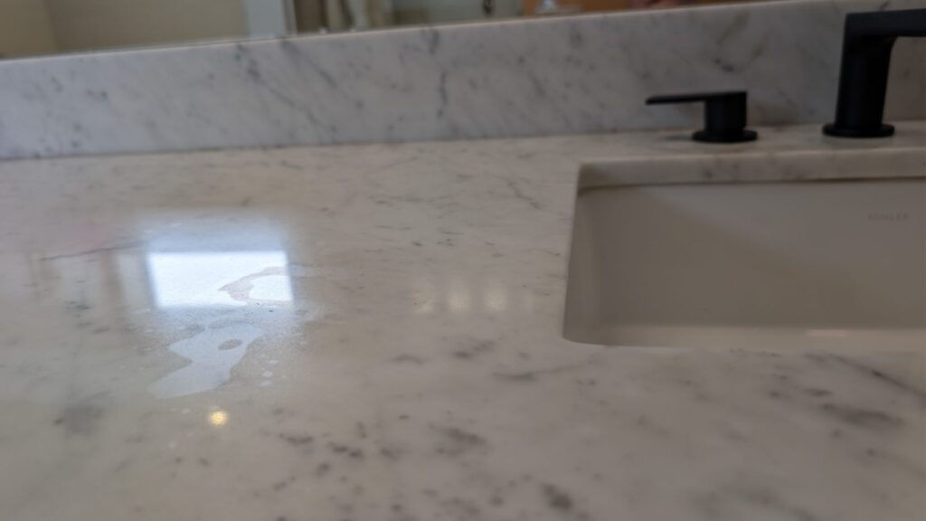 Etch mark on a Carrara marble bathroom vanity beside a black faucet, before restoration