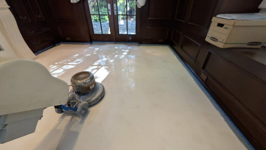 A floor machine working across a pale limestone floor beside dark wood cabinetry taped off with blue painter's tape, in a Point Grey home