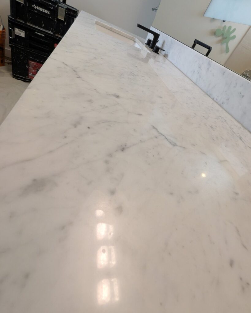 White marble vanity top seen along its length, hazy with etch marks and a blurred reflection of the lights, before restoration