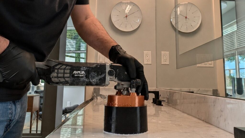 Gloved hands guiding a handheld wet polisher across a white marble vanity top, the slurry visible on the stone